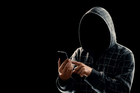 Silhouette of a man in a hood on a black background, his face is not visible, the hacker is holding the phone in his hands. The concept of a criminal, incognito, mystery, secrecy, anonymity.の写真素材