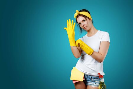 A girl, a cleaning woman before cleaning, is putting on her hands rubber, yellow gloves on a blue background. The concept of cleanliness in the house, cleaning of the premisesの写真素材