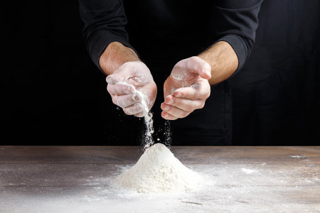 Hands of the cook close-up, pour the flour on the table to cook dough on a black background.