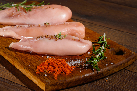 Raw chicken breast, pepper, rosemary on a cutting board, on a wooden background, close-up. Raw chicken meat for cooking. Delicious balanced nutrition concept. Copy spaceの写真素材
