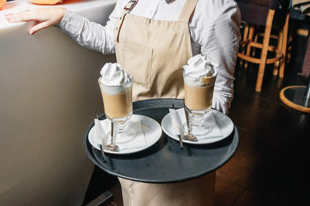 Cold coffee with whipped milk and caramel ice cream in high glasses on a tray in the hands of the waiter, selective focusの写真素材
