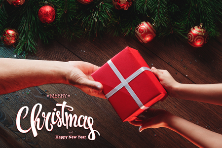 The inscription of Merry Christmas and Happy New Year, male hands hold a gift on a wooden brown table. Christmas card, holiday background. Mixed media. View from above.の写真素材