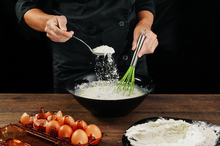 Male hands chef pours the flour into a plate on a wooden brown table in black utensils. Chef kneads the dough. Black background. The concept of cooking food. Close-up. Copy space.の写真素材