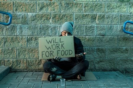 A man, homeless, a man asks for alms on the street with a sign will work for food. Concept of homeless person, addict, poverty, despairの写真素材