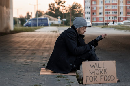 A man, homeless, a man asks for alms on the street with a sign will work for food. Concept of a homeless person, social problem, addict, poverty, despairの写真素材