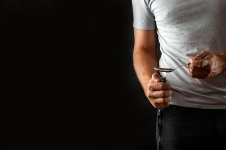 A male drug addict is preparing drugs in a spoon with a lighter on a black background. Social...