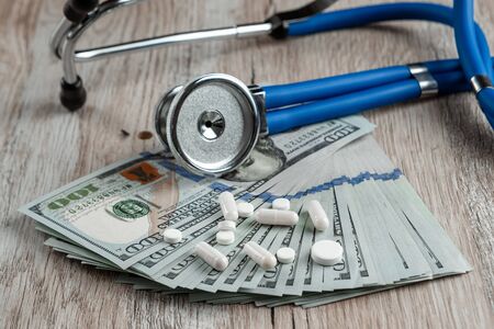 A stethoscope lies on a bundle of money and pills on a light wooden background. The concept of expensive medicine, medical insurance, expensive drugs. Copy spaceの写真素材