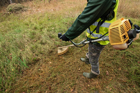 A working man in professional outfit mows grass with a trimmer, a mower. Mowing lawns, roadsides,...