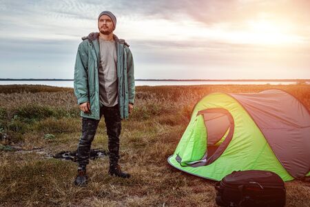 A bearded man near a camping tent in green on the background of nature and the lake. The concept of travel, tourism, campingの写真素材