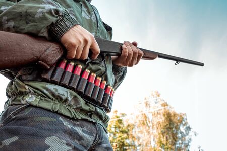 A hunter with a gun in his hands in hunting clothes in the autumn forest close-up. The hunting period, the fall season is open, the search for preyの写真素材