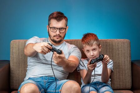 A cheerful family, dad and son play on the console, in video games, emotionally react while sitting on the couch. Day off, entertainment, leisure, spend time togetherの写真素材