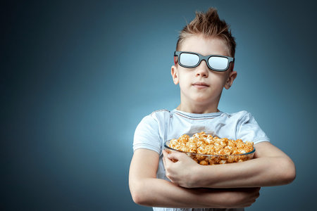 The boy holds popcorn in his hands watching a movie in 3D glasses on a blue background. The concept...