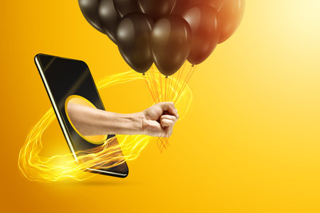 Hand holds a bunch of black balloons through a smartphone screen on a yellow background. Holiday...