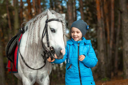 Little girl portrait, stands next to a white pony close-up on the background of nature. Jockey, epodrome, horseback riding.の写真素材