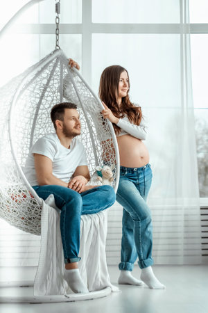 A pregnant girl with her husband are standing in a bright bedroom. Beautiful belly of a young attractive pregnant girl. Family, marriage, childbirth conceptの写真素材