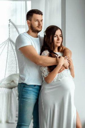 A pregnant girl with her husband are standing in a bright bedroom. Beautiful belly of a young attractive pregnant girl. Family, marriage, childbirth conceptの写真素材