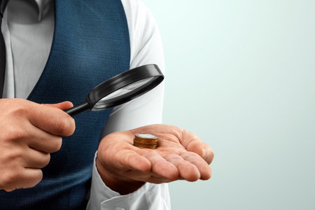 A man looks through a magnifying glass at a stack of coins in his palm. Small salary, financial crisis, bankruptcyの写真素材