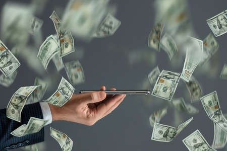 The hand of a businessman holds a smartphone on the background of falling dollars. The concept of money transfers, internet banking, online work, sports bettingの写真素材