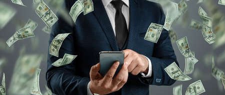 Online sports betting. A man in a suit is holding a smartphone and dollars are falling from the sky. Creative background, gamblingの写真素材