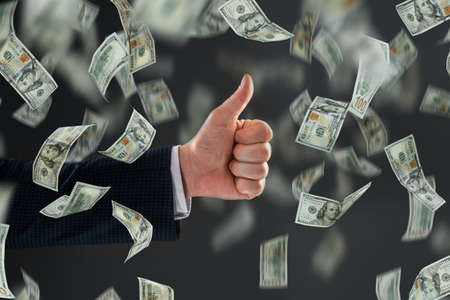 Businessman's hand shows thumbs up against the background of falling dollars. The concept of investments, dividends, interest, bank depositsの写真素材