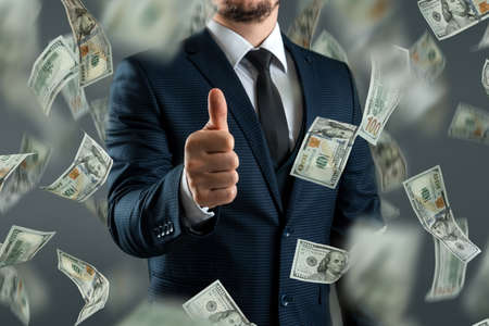 A businessman in a suit shows thumbs up against the background of falling dollars. The concept of investments, dividends, interest, bank depositsの写真素材