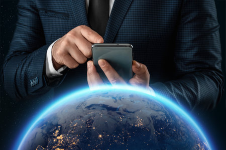 A man in a business suit, a businessman holds a smartphone in front of the globe. Mixed media. Concept Global networks and international business.の写真素材