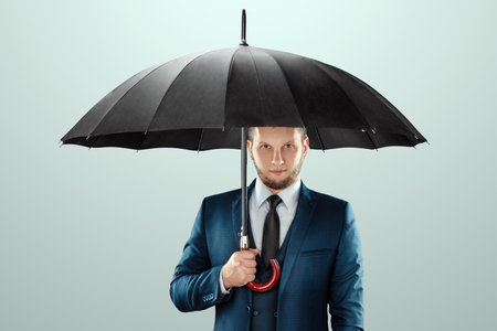 A man in a business suit stands with an umbrella in his hands on a light background. Savings and assets protection, insurance conceptの写真素材