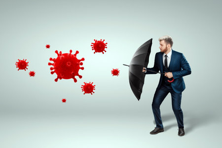 A man in a business suit stands with an umbrella in his hands and protects his business from the coronavirus. COVID-19 concept, protection, savings and assets insuranceの写真素材
