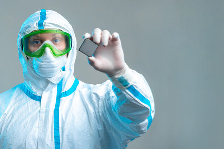An employee holds a microchip in his hands, stands in a protective suit, glasses, a mask and gloves....