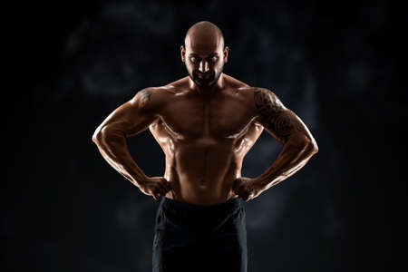 Male bodybuilder with light stubble and bare torso shows muscularity against a dark background. The concept of a fitness club, doing sports, weightlifting. Copy spaceの写真素材