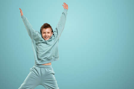 Little in a turquoise suit poses and fools around on a turquoise background, looks at the camera. Children's studio portrait. Childhood lifestyle conceptの写真素材