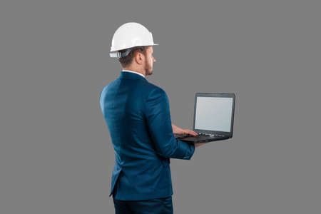 A man in a business suit, an architect with a white helmet on his head, holds a laptop, isolated on a white background. Architecture, construction conceptの写真素材