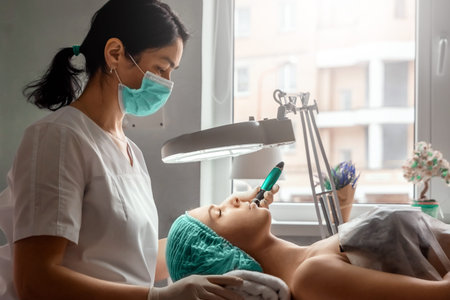 A beautician wearing medical gloves cleans the pores on the face using special equipment while...