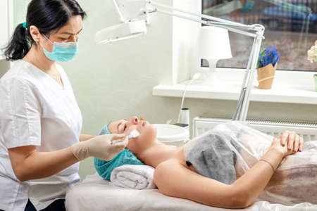 A beautician applies a mask to the face in a beauty salon. The concept of facial hygiene, cosmetic procedures, skin care, skin pore cleansing, hardware cosmetologyの写真素材