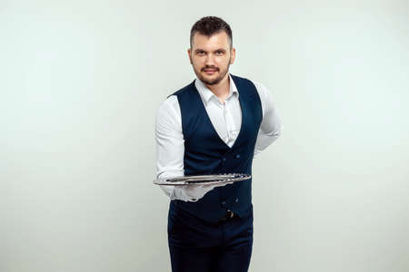 Handsome male waiter, white shirt, holds a silver tray, hand behind his back. Concept of wait staff serving customers in a restaurantの写真素材