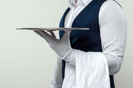 A male waiter in a white shirt and white gloves stands sideways with a silver tray. The concept of service personnel serving customers in a restaurantの写真素材