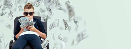 A cute, rich boy with glasses holds dollars and knows how to make money against the background of falling banknotes. Money rain, Financial education, young millionaire, investments, loans, winningsの写真素材