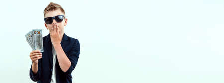 A rich boy on a light background holds a bundle of dollars in his hands. Financial education, young millionaire, investments, loans, winnings, dividends. Flyer blank, site header, copy spaceの写真素材