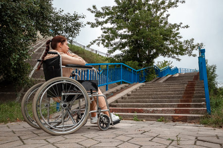 A young disabled girl sits in a wheelchair in front of the stairs. The concept of a wheelchair, disabled person, full life, paralyzed, disabled person, health care, lonelinessの写真素材