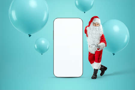 Smartphone mockup image, environment to showcase mobile app design, creative background. Smartphone for your advertising. Santa claus is standing next to a large smartphone. Christmas advertisementの写真素材