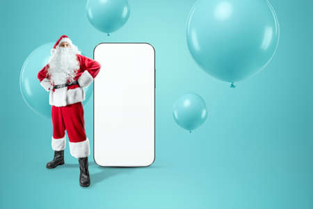 Smartphone mockup image, environment to showcase mobile app design, creative background. Smartphone for your advertising. Santa claus is standing next to a large smartphone. Christmas advertisementの写真素材
