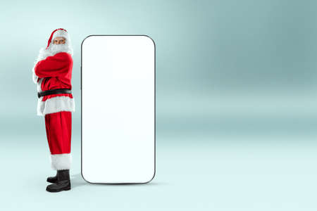 Smartphone mockup image, environment to showcase mobile app design, creative background. Smartphone for your advertising. Santa claus is standing next to a large smartphone. Christmas advertisementの写真素材