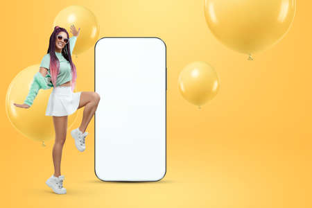 A creative young girl stands near a large smartphone with a white screen. Copy space, mixed media, ad layout, empty design environment, creative backgroundの写真素材
