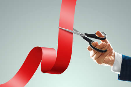 Grand opening with red ribbon and scissors. A businessman's hand holds scissors cuts a red ribbon on a light background. Close up, copy spaceの写真素材