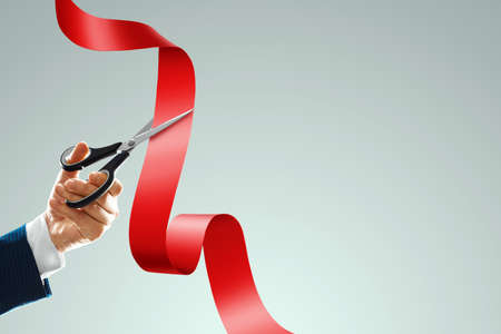 Grand opening with red ribbon and scissors. A businessman's hand holds scissors cuts a red ribbon on a light background. Close up, copy spaceの写真素材