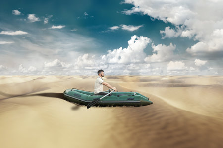 A man sits in an inflatable boat in the desert, yellow sand and blue sky. Problem concept, drought, melting glaciers, global warming, crisis, depression. mixed media, copy spaceの写真素材
