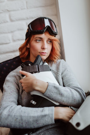 Woman holds white ski boots and looks out the window because she cannot go skiing because of a...
