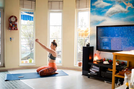 Beautiful woman stretching her arms while doing yoga in her cozy apartmentの写真素材