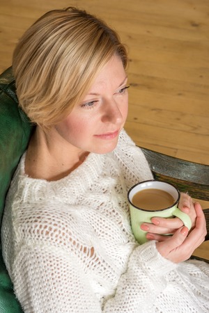 portrait of a woman in a white sweater, which sits in a leather chair and drinking coffee with milkの写真素材