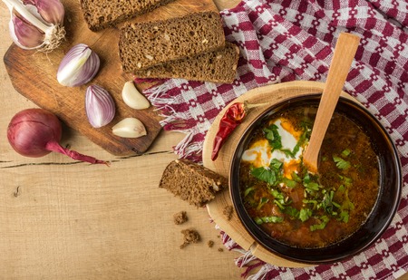 Tasty and nutritious borsch-soup with meat, potatoes, cabbage, tomatoes, beans, sour cream, parsley, onion, garlic, dark bread in a clay plate on a wooden tray and wooden table. Top viewの写真素材